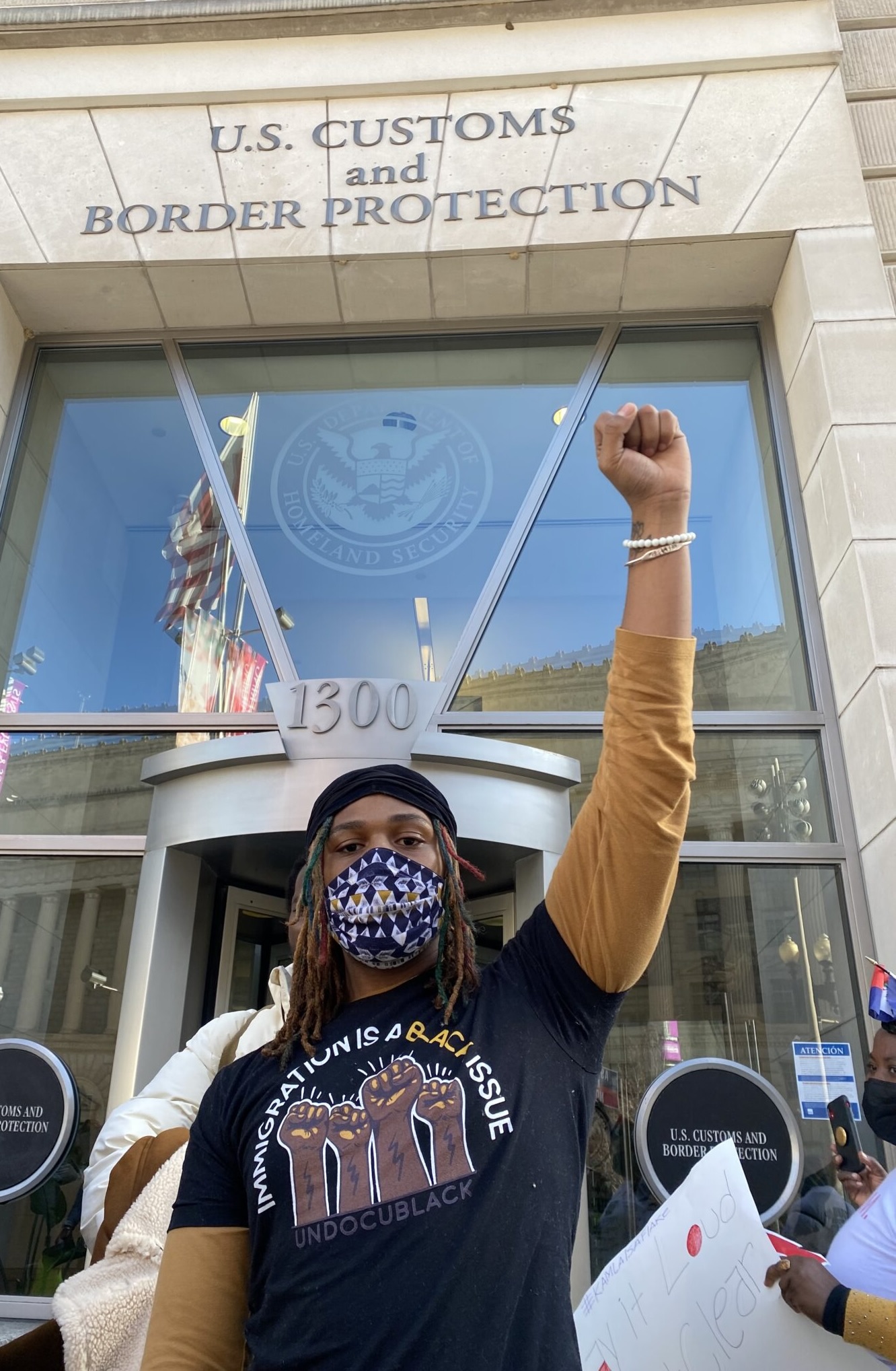 Masked person in a t-shirt that says "Immigration is a Black Issue" looking at the camera with a raised left fist outside of a U.S. Customs and Border Protection office