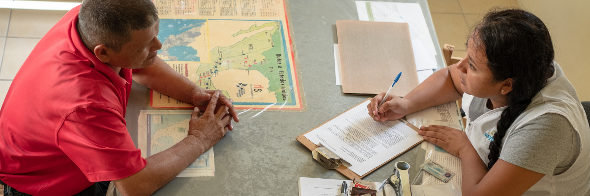 A man sitting at a table with a map in front of a woman with a clipboard and a vest