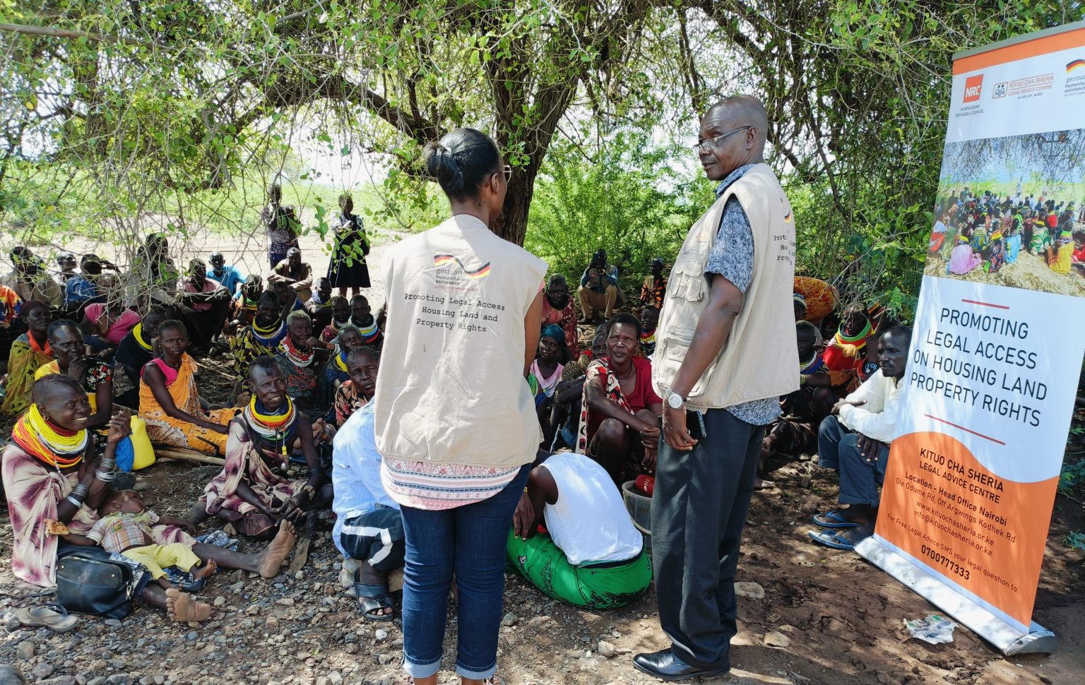 Two staff members in beige vests talking about promoting legal access on housing, land, and property rights to a group of people sitting on the ground 