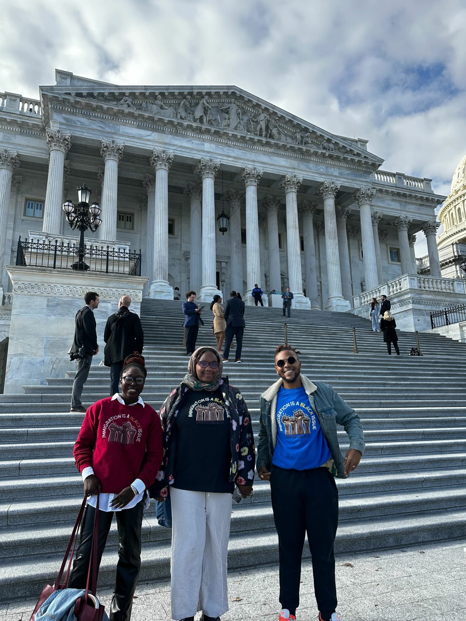 Three people standing outside at the bottom of the Supreme Court steps wearing shirts that say "Immigration is a Black Issue"