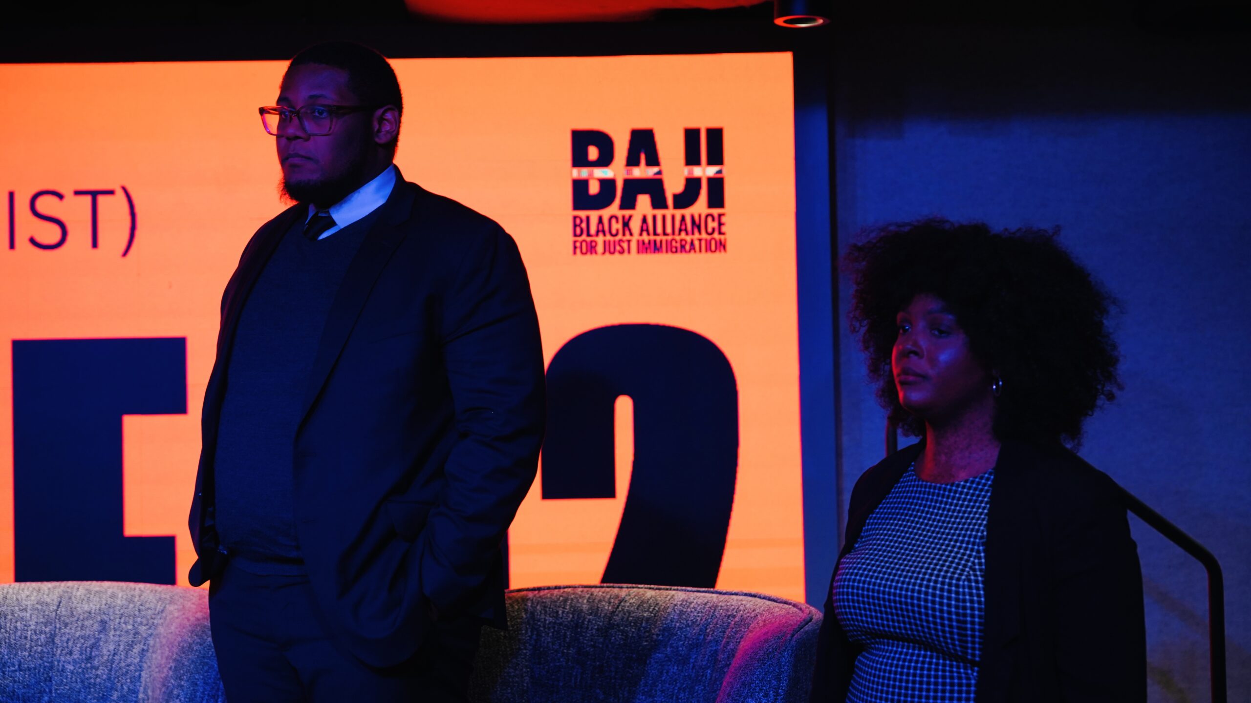 One man is standing and a woman is sitting in front of a banner at a gala 